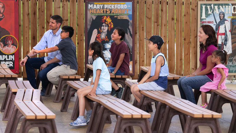 Mixed group of adults and children sit on benches at Boomtown 1905's outdoor Majestic Theatre. Vintage film posters are shown in the background, attached to a wooden fence post. 